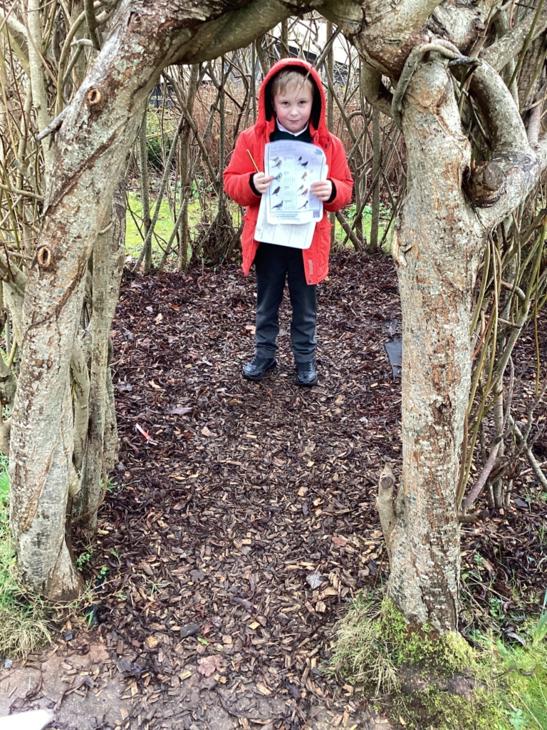 Exwick Height Pupil stood between two tress in red coat holding a bird watch survey. 