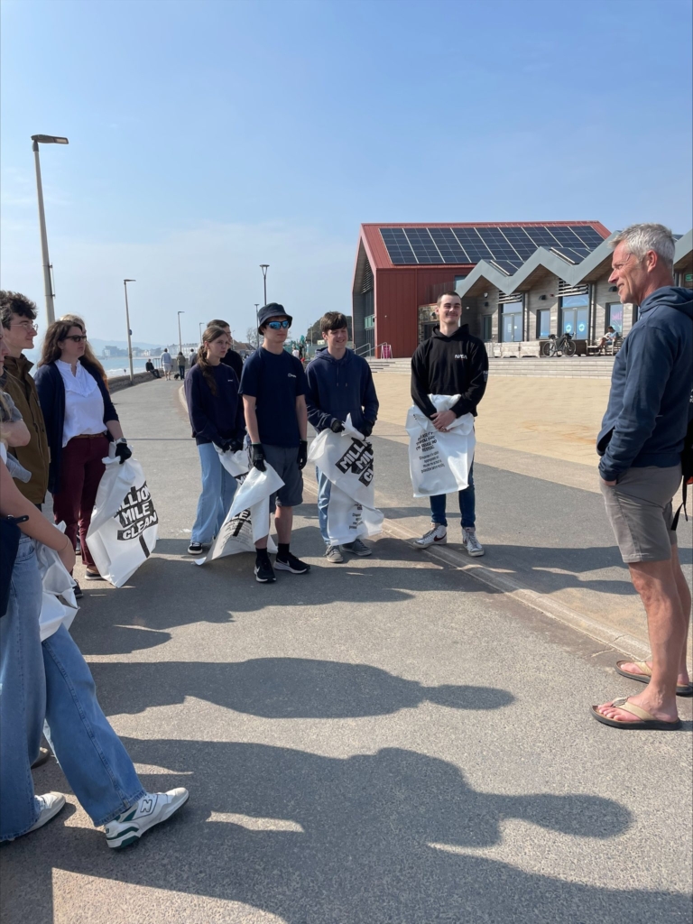 Student stood in semi circle listening to speaker at seafront