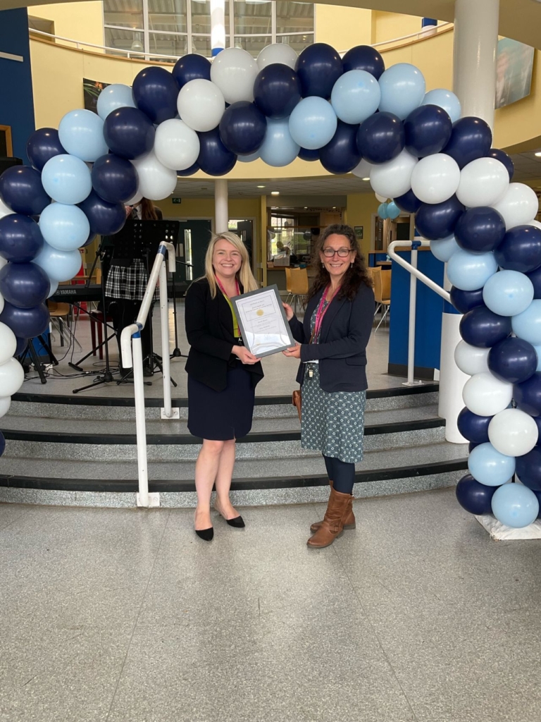 Two staff members stood posing for the camera in front of a blue balloon arch receiving their award