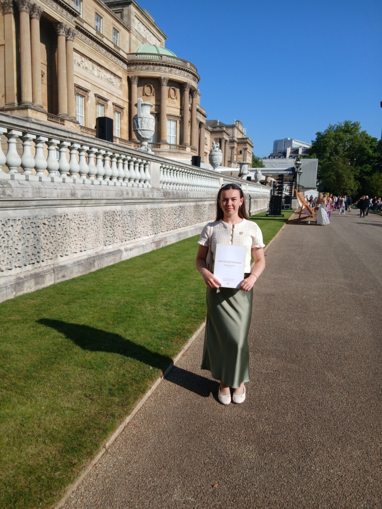 Young women stood in front of a green mowed law holding a certificate, outside of Buckingham Palace