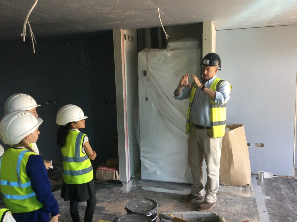 Children stood in hard hats and high vis jackets in a room under construction