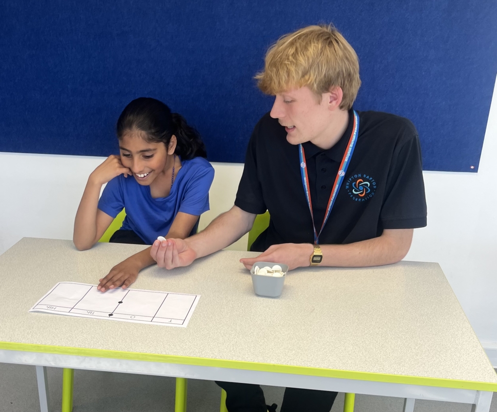 Member of staff from Whipton Barton Federation sat at a table teaching a pupil. 