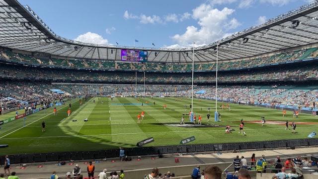 Photo of a rugby stadium. In the distance you can see thousands of seats filled with people watching the game, in the foreground you can see the green rugby pitch with players.