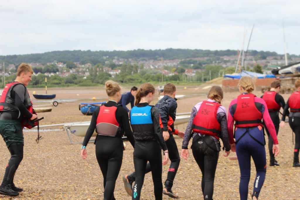 Photo of the backs of students in wetsuits and life jackets walking along the beach