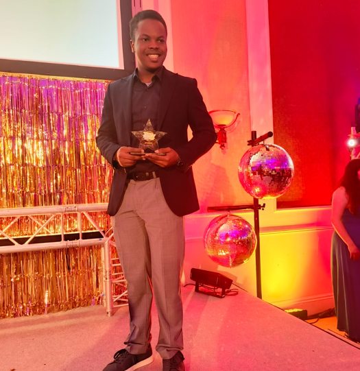 Student stood on stage in a suit, holding award and smiling at the camera