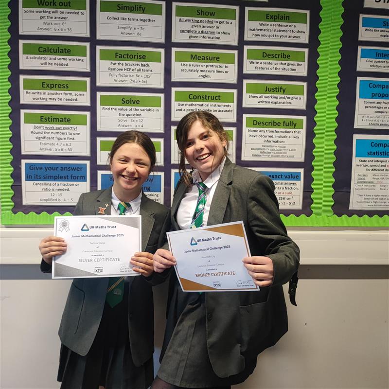 Two students in uniform holding up certificates smiling, in front of wall display in school corridor