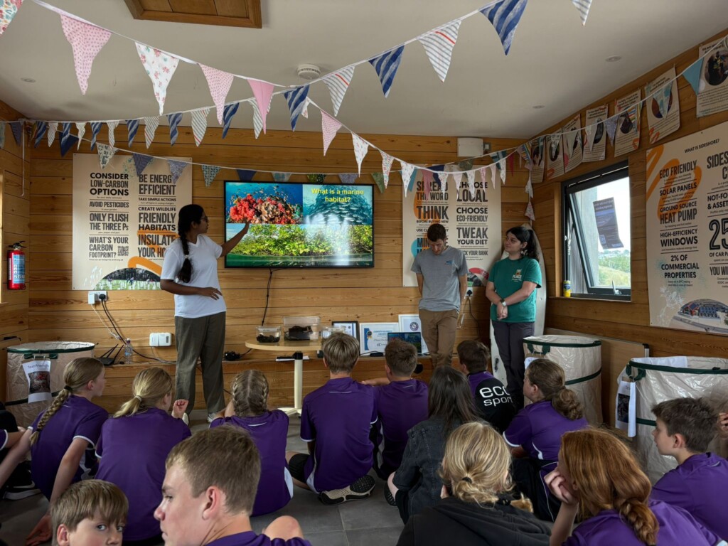 Students sat on the floor with their backs to the camera watching a presentation in a wooden hut