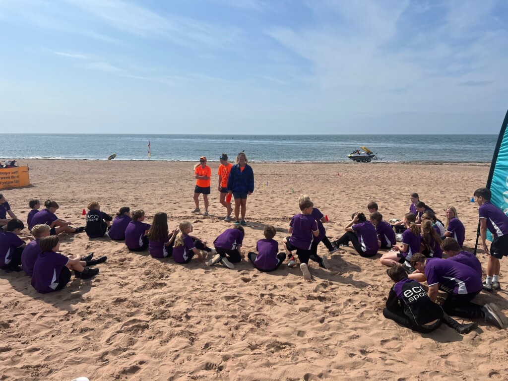 Students sat in a line with their backs to the camera, on the sand facing the sea