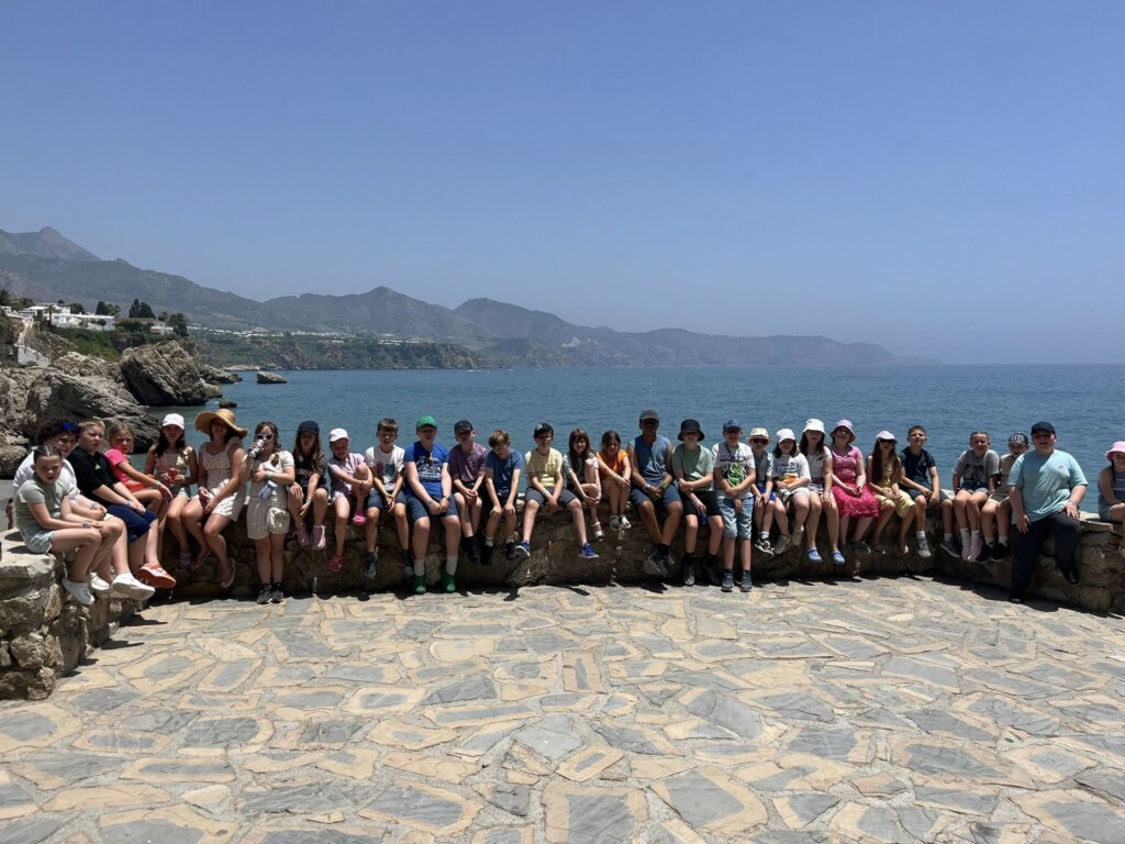Students sat in a line on a sea wall facing the camera. In the distance you can see the sea and hills