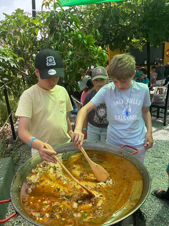 Two students stirring a big pan of paella in a garden