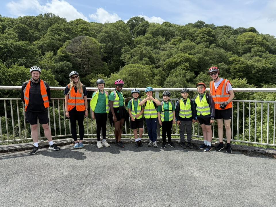 Young students stood in front of railing in high vis jackets and helmets. Green trees in background.