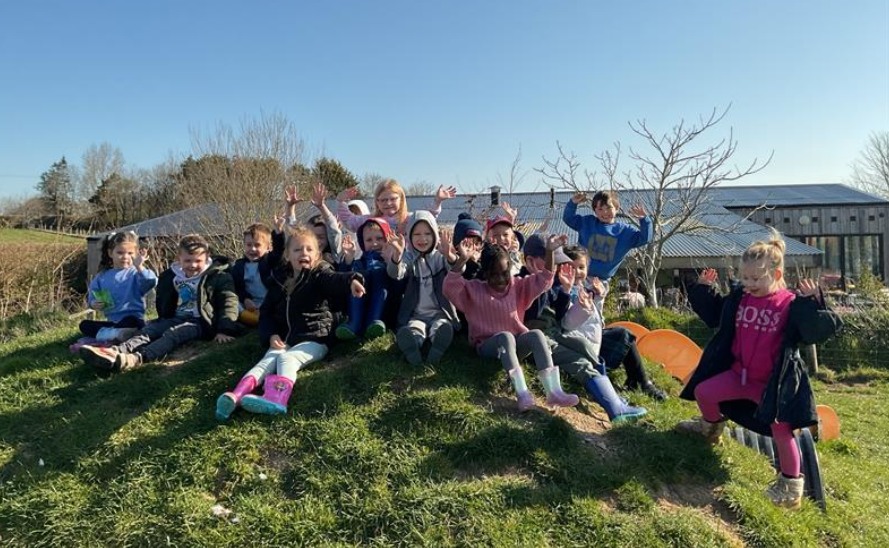 Students sat on green grass, smiling and waving at camera