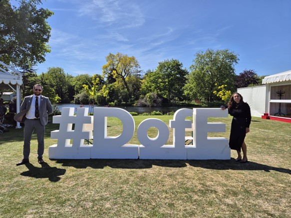 A large white DofE sign with two people stood either end of the sign, on grass