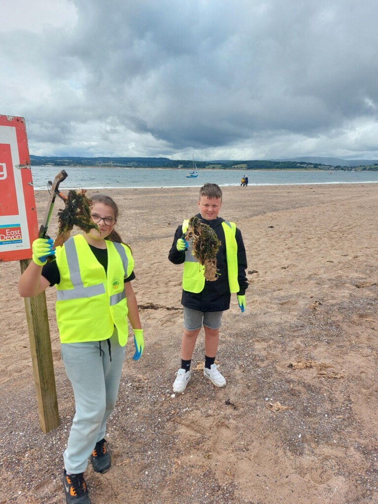 Two students stood on the beach in high vis yellow jackets, picking up rubbish