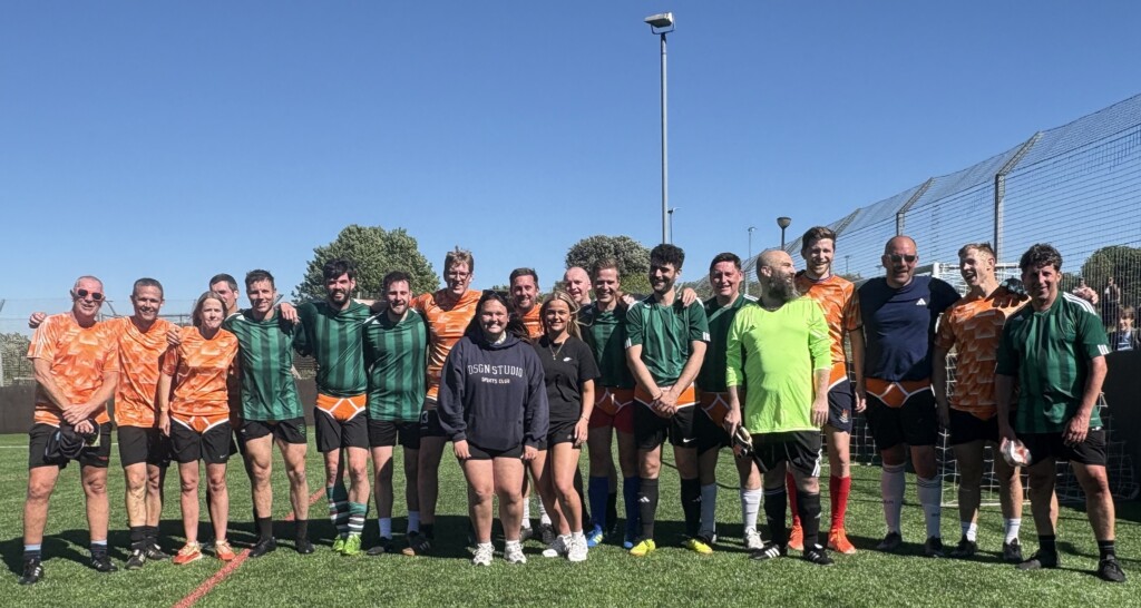 Group of staff in football kits stood in a line facing towards the camera, stood on grass