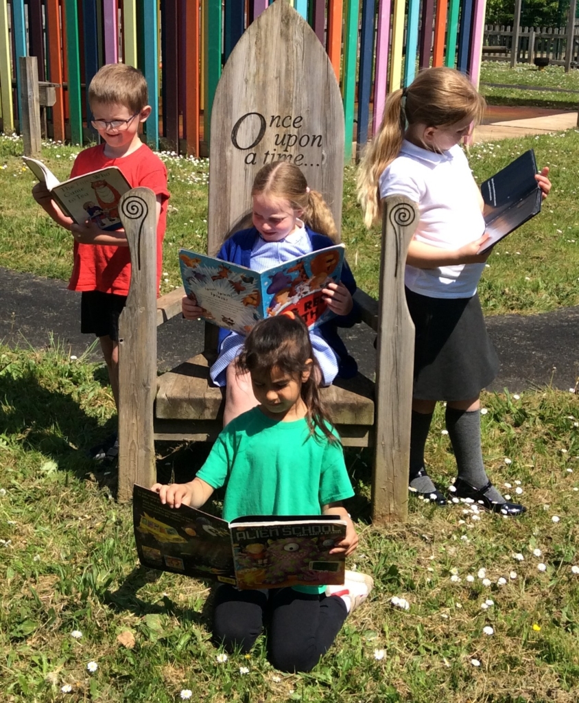 One child sat on a wood chair reading, one child sat ion front of the chair on the grass reading and one child stood either side of the chair reading