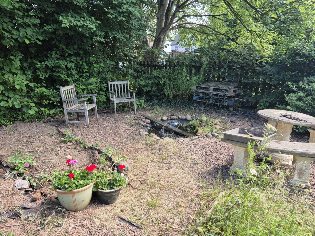 A section of the school garden bounded by a fence. Dirt ground with pots of flowers dotted around and benches at the far end against the fence.