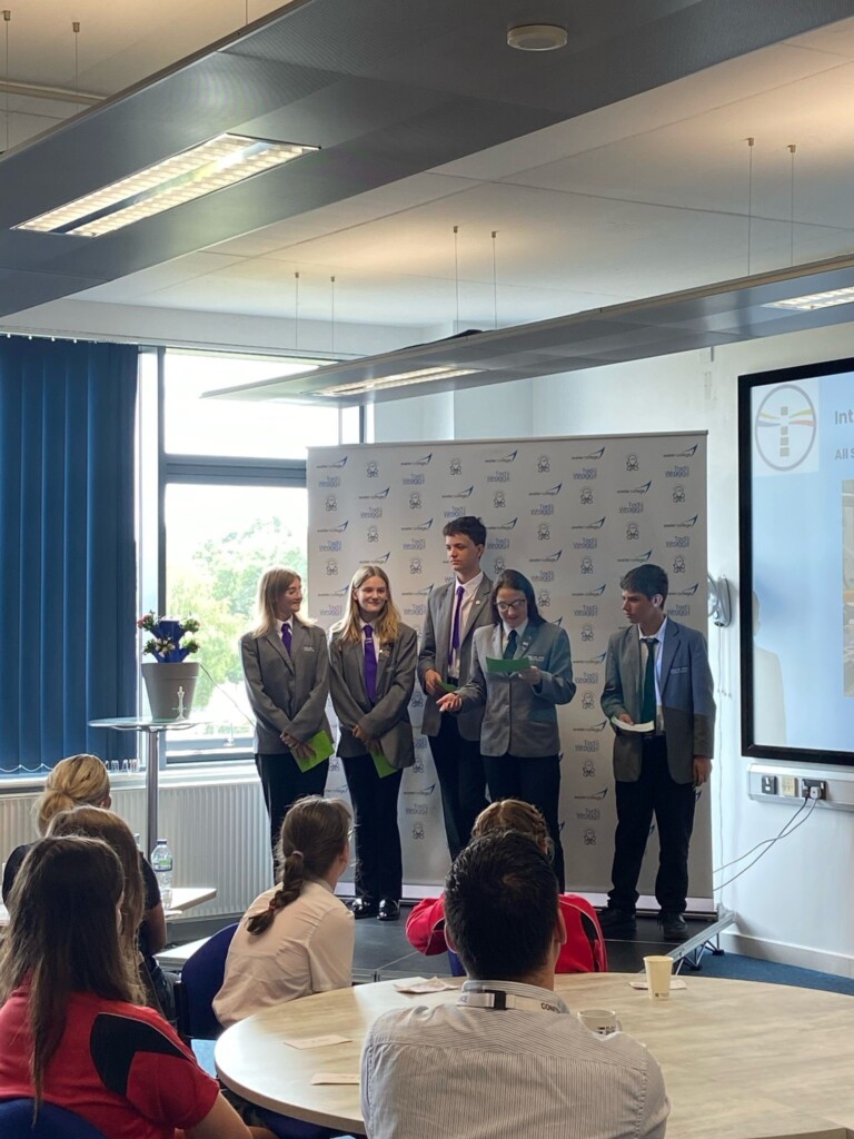 Five students in grey blazers stood presenting to an audience on a small stage