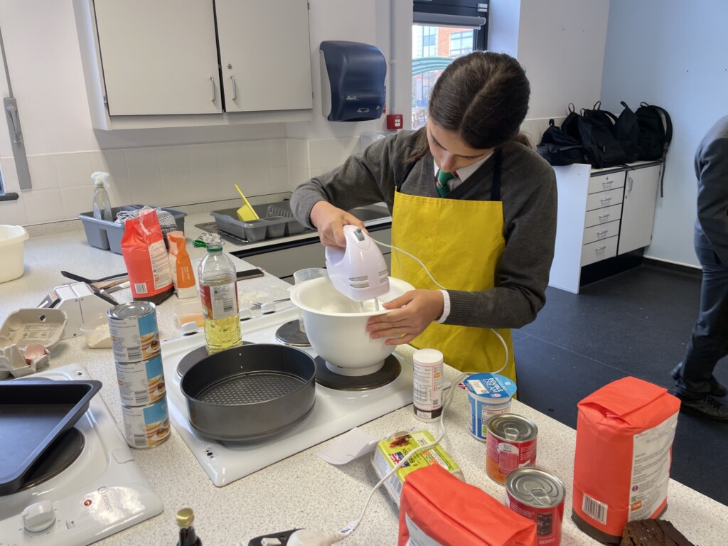 One student in yellow apron whisking cake batter in a bowl. Stood in white kitchen.