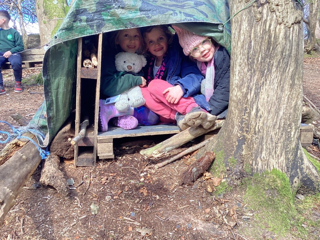 Three students sat under a green plastic sheet in a makeshift den in the woods