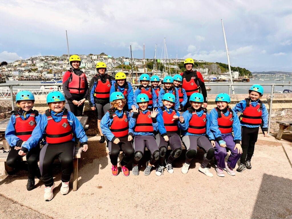 Group of students in blue helmets, red life jackets and wetsuits posing for photo by the sea