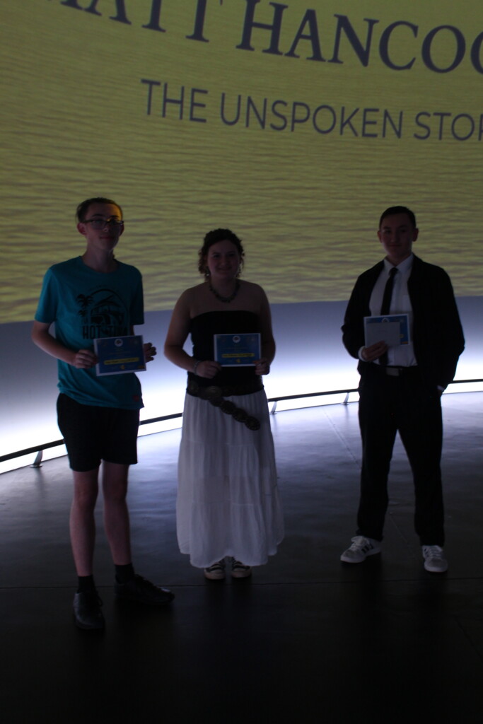 Three students stood in front of screen holding certificates