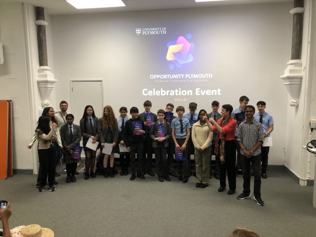 Two rows of students stood facing camera, in a classroom in front of a presentation screen