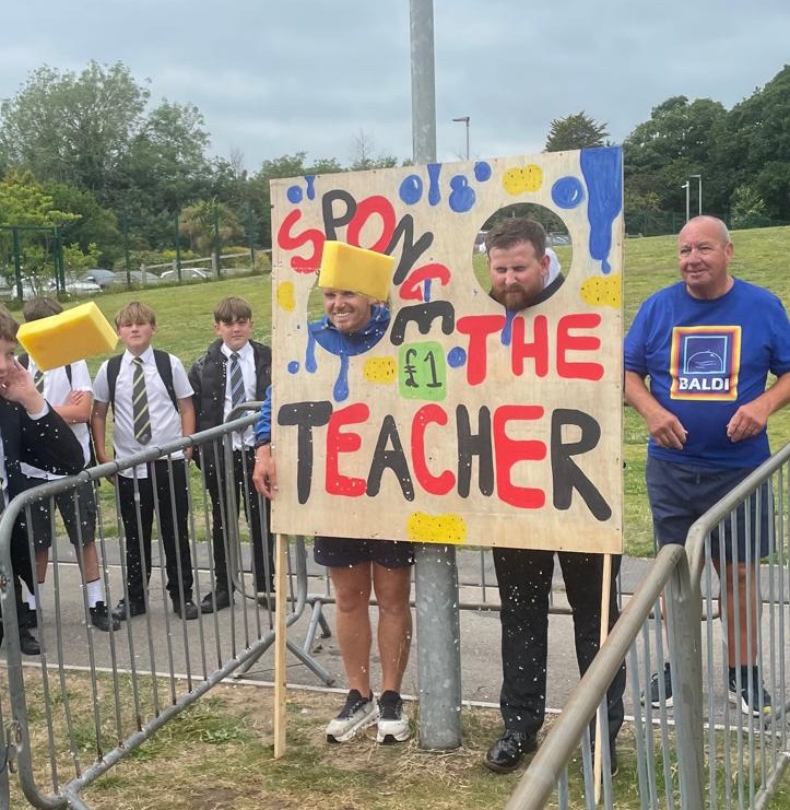 Two teachers stood with heads poking through holes in a square of wood with sponge the teacher written across it