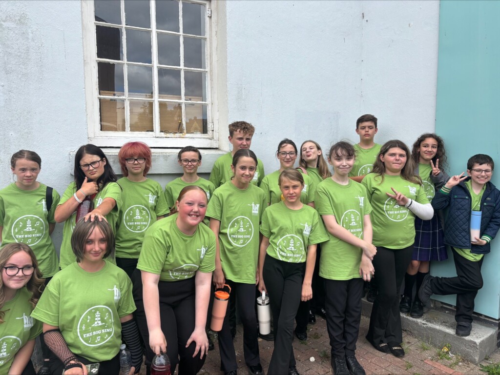 Two rows of students stood outside building smiling to the camera, all wearing green t-shirts.