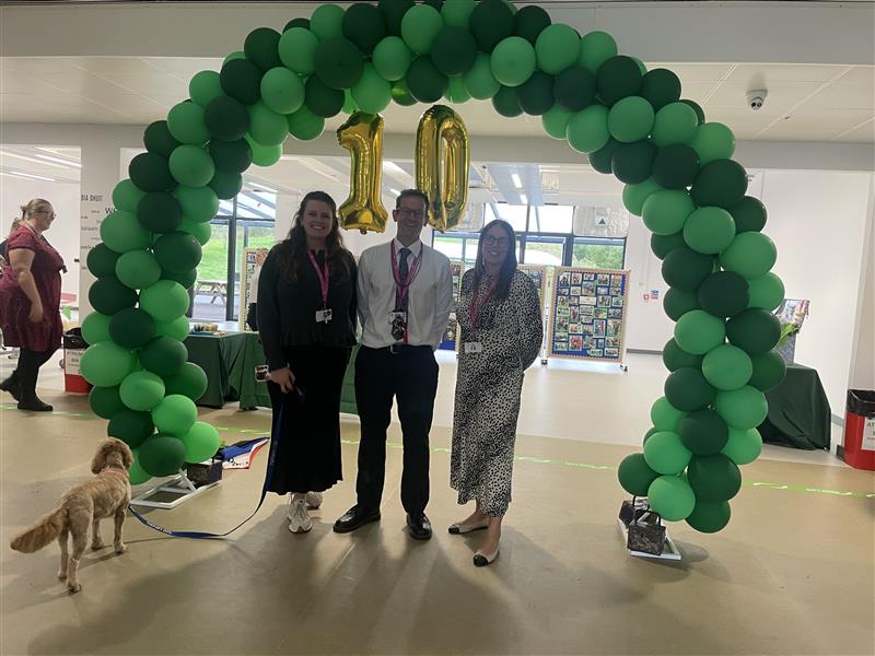 Three members of staff stood in front of green balloon arch with the number 10 spelled out by balloons