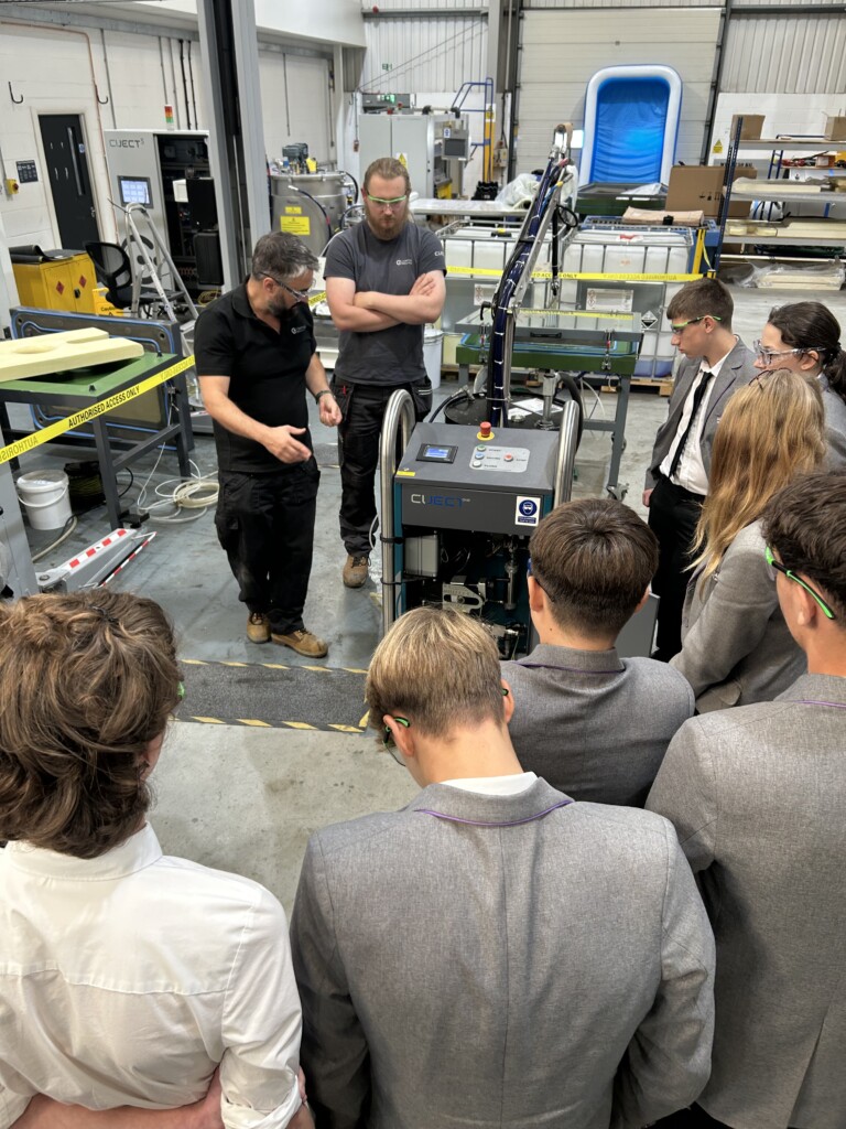 A group of students in grey blazers gathered around a large machine during a workshop
