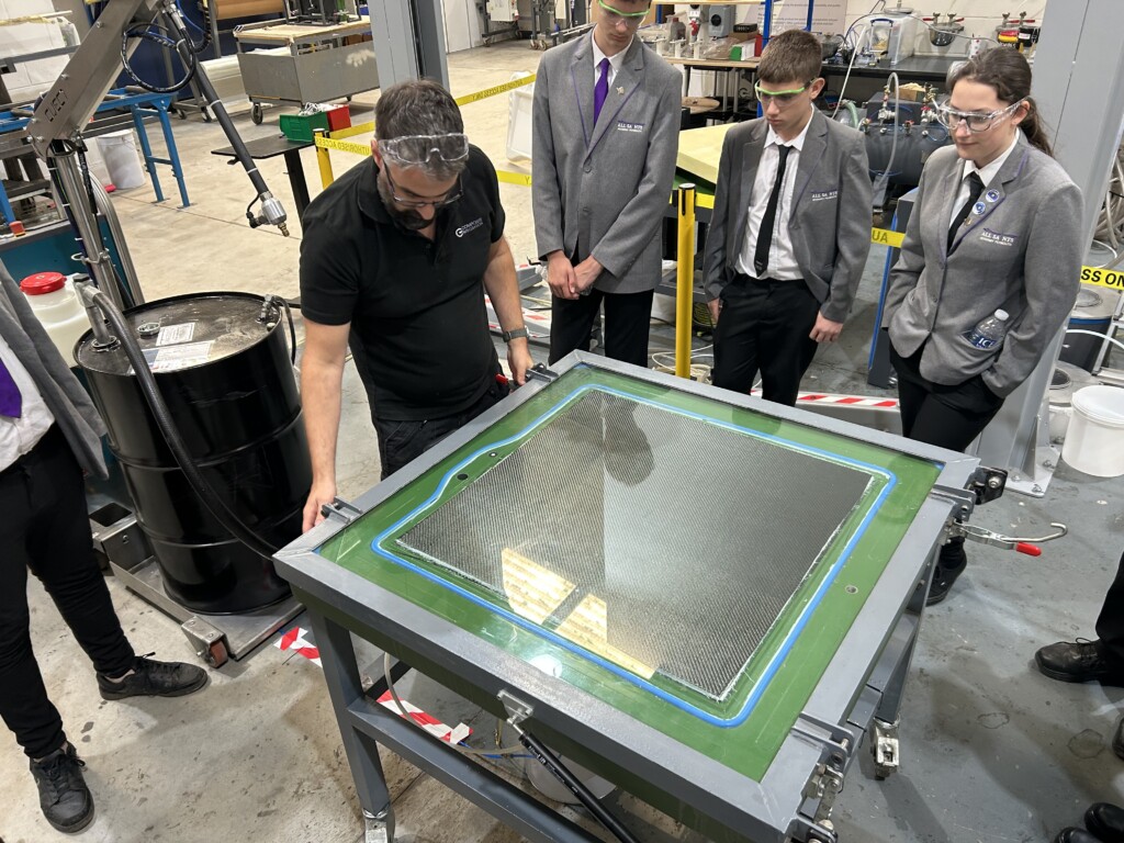 A couple of students watching an engineer use a piece of machinery in the shape of a square table