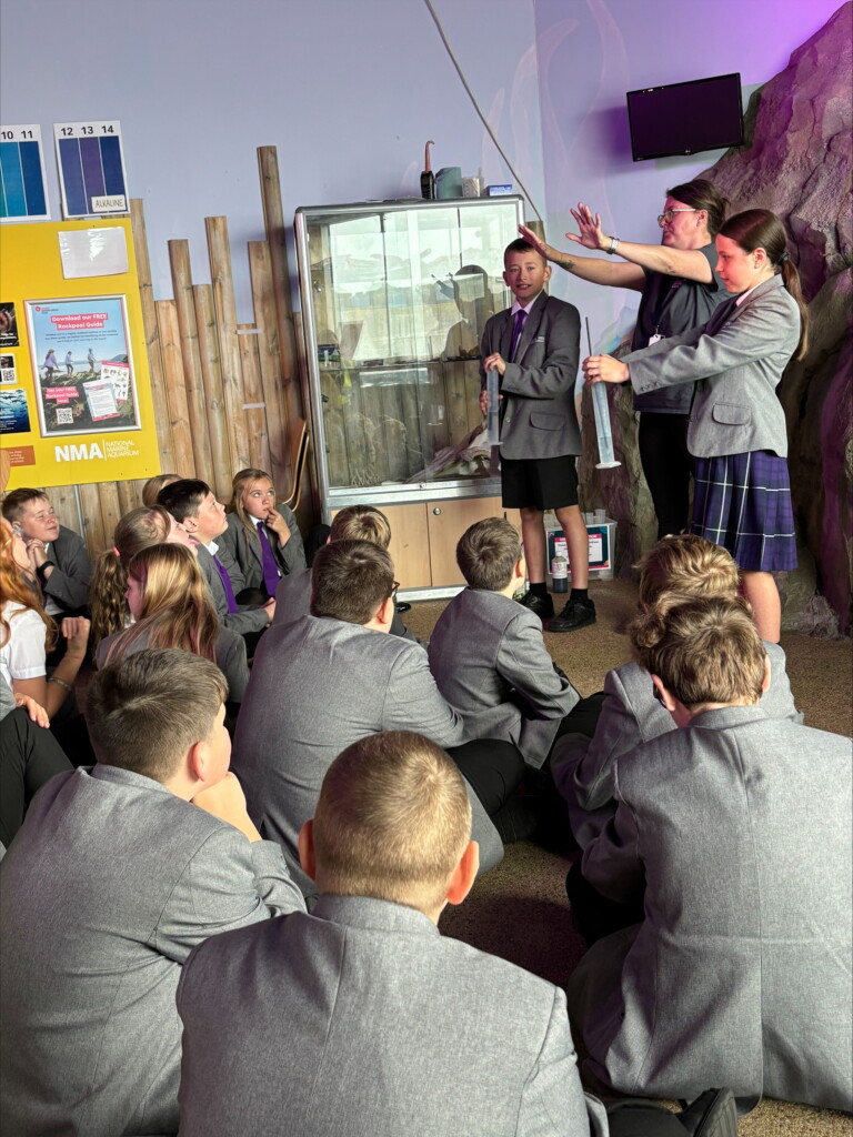 Students sat on floor watching a presentation with two student stood at the front part of a demonstration