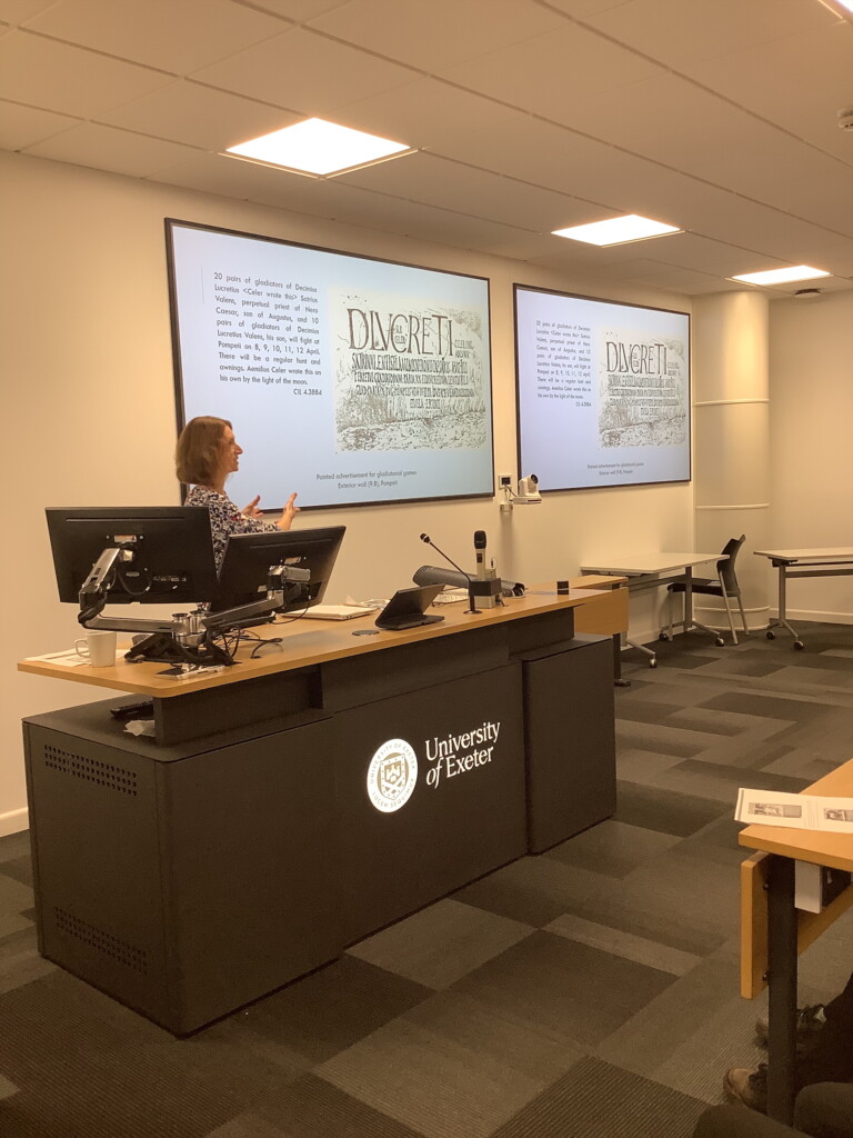 A black desk with a teacher stood up behind it giving a lecture