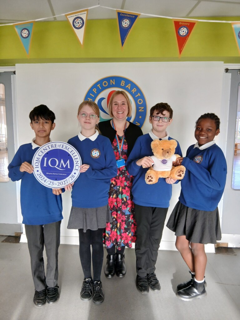 Students and a teacher have a picture taken with the IQM Award and a teddy.