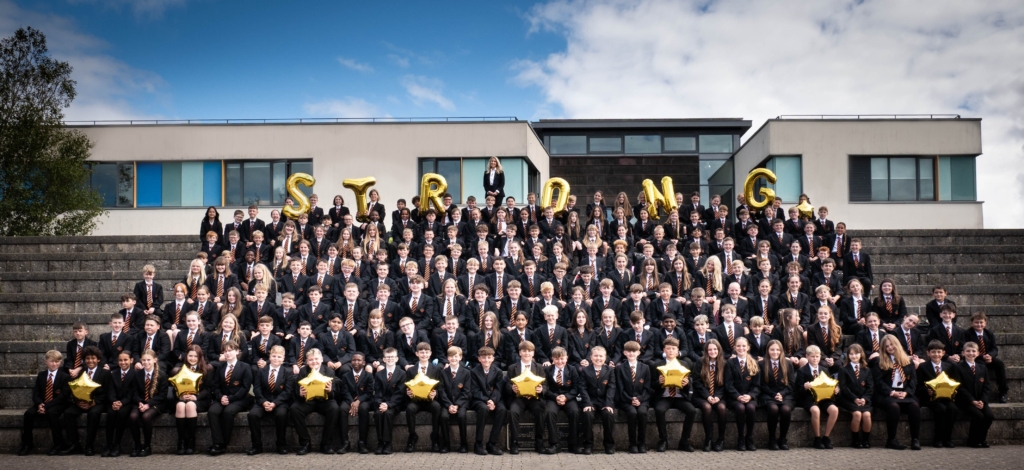 A group of students sat in rows leading up stairs, with balloons spelling STRONG along the top and headteacher Tracy in the middle